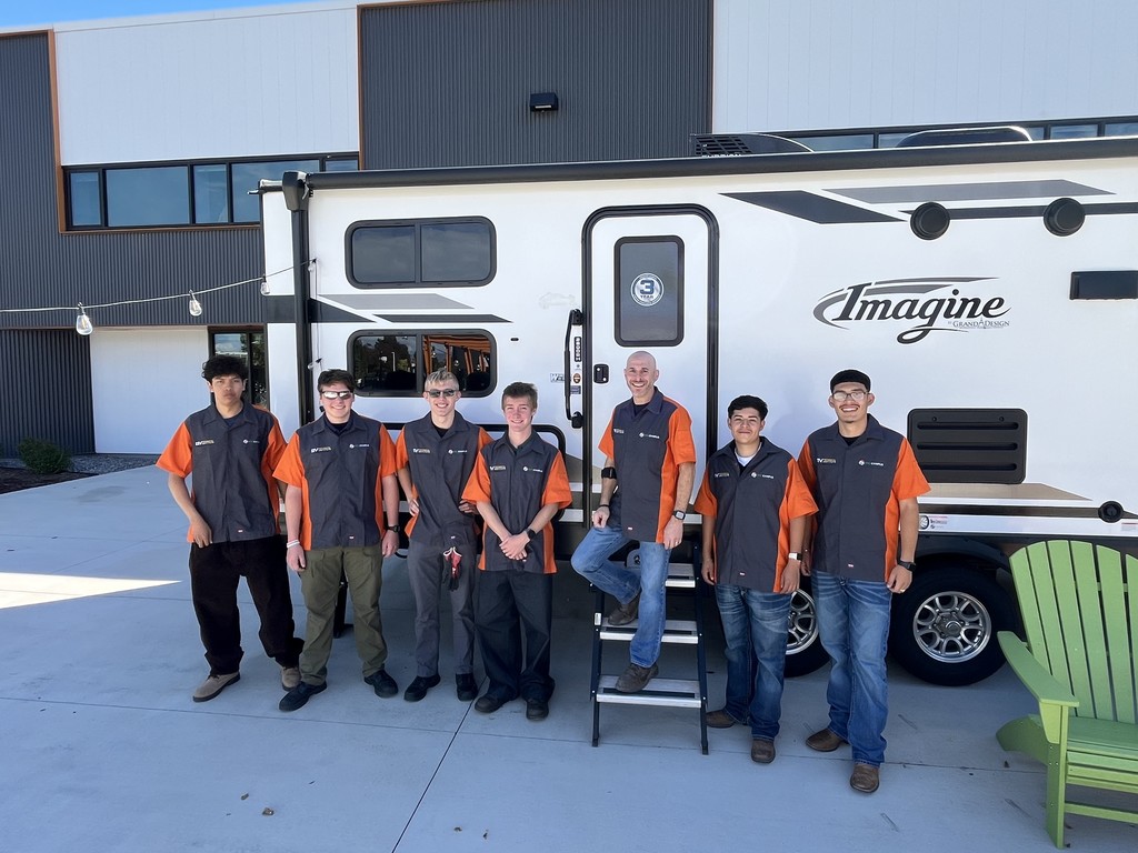 Eight individuals stand in front of a white "Imagine" Grand Design RV parked outside a modern industrial-style building. They are wearing matching uniforms with dark gray vests and bright orange short sleeves, suggesting they are part of a team or program. One adult stands on the RV steps, while the rest line up beside him. The group appears to be male and includes both youth and adults. The background features string lights, a green Adirondack chair, and a concrete courtyard.