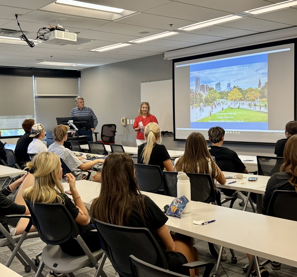 Classroom scene with a group of high school or college students seated at tables, attentively listening to two adult presenters—one man standing at a podium and one woman in a red blouse speaking near a projection screen. The screen displays a presentation titled “Career Paths, Companies in Colorado and Core Competencies for Real Estate” with an image of an urban park and city skyline. The room is modern, with white walls, ceiling lights, and a ceiling-mounted projector. Students are diverse in appearance and are casually dressed.