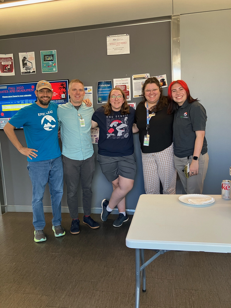 Five staff members pose for a group photo indoors in front of a wall filled with colorful posters and event flyers. They are smiling and standing close together, showing camaraderie. One wears a blue EPIC Campus t-shirt, another a MSU Denver shirt, and others wear staff badges or branded polos. A folding table with paper plates and a Diet Coke can is in the foreground.