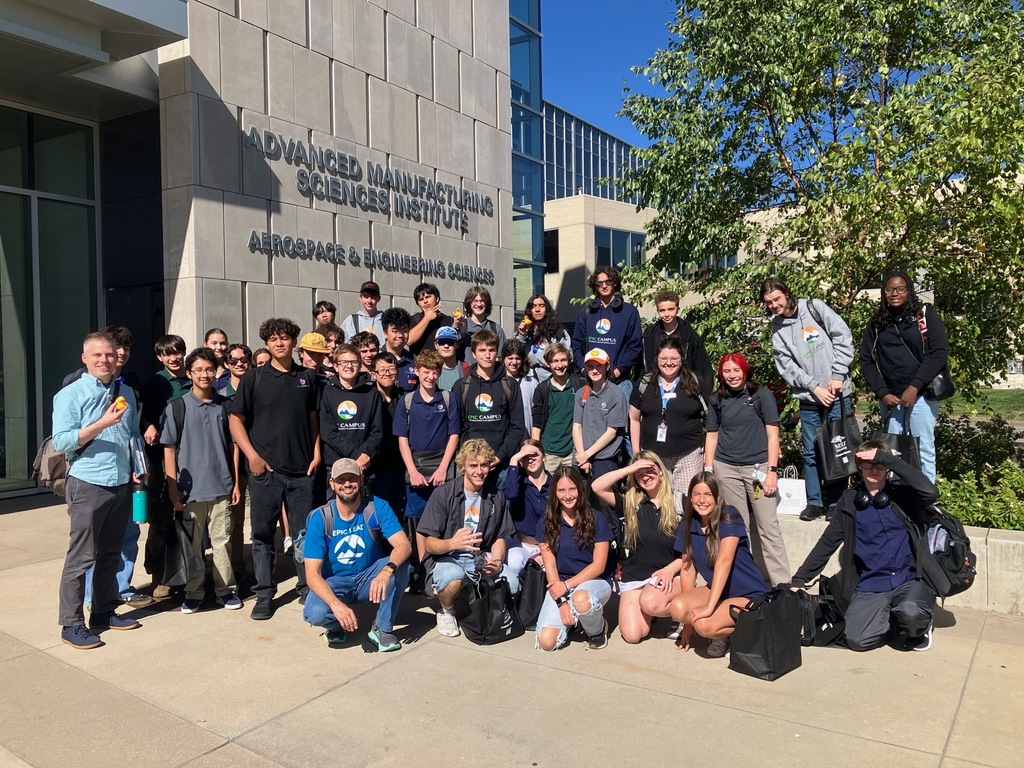 A large, diverse group of high school students and a few adults pose in front of the "Advanced Manufacturing Sciences Institute" building at MSU Denver. The signage also reads "Aerospace & Engineering Sciences." Most students wear EPIC Campus gear, and several are smiling or making peace signs. The weather is sunny with a clear blue sky, and the group stands on a sidewalk beside a modern building and a leafy tree.