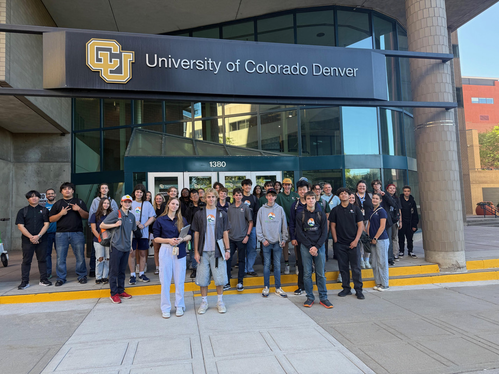 A large group of high school students stands outside the University of Colorado Denver, under a large black sign with gold and white CU branding. The address "1380" is visible above the glass entrance doors. Students are smiling, wearing a mix of EPIC Campus shirts and casual clothing. The scene is sunny, with shadows cast on the pavement, suggesting a campus tour or visit.