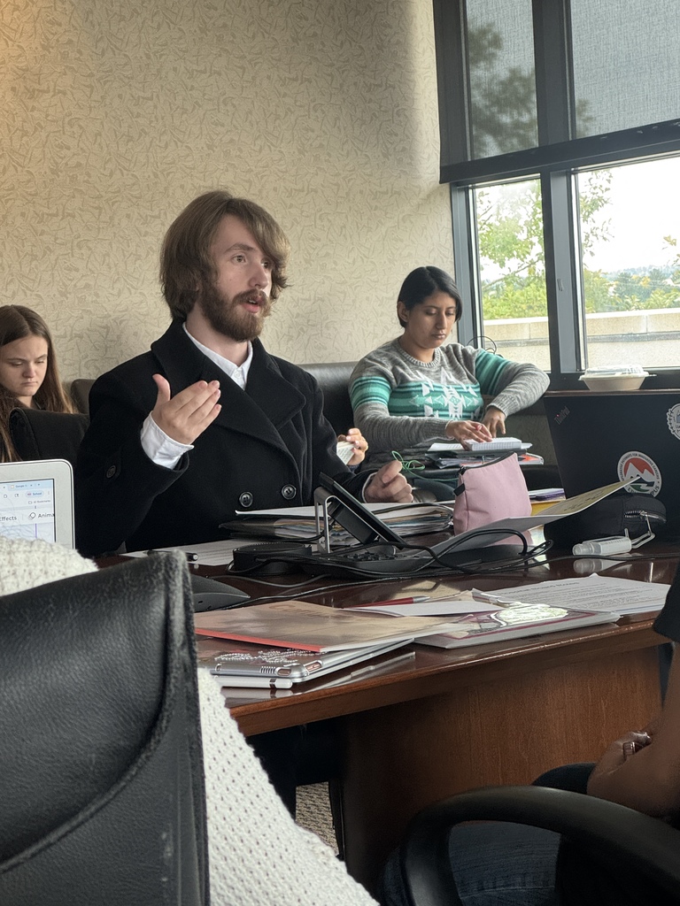 A young man in a black peacoat sits at a large conference table, speaking and gesturing with one hand while surrounded by notebooks, folders, and a laptop. Other students sit nearby, including a girl in a striped sweater taking notes. The setting is a meeting room with large windows and natural light. The atmosphere is focused and academic.
