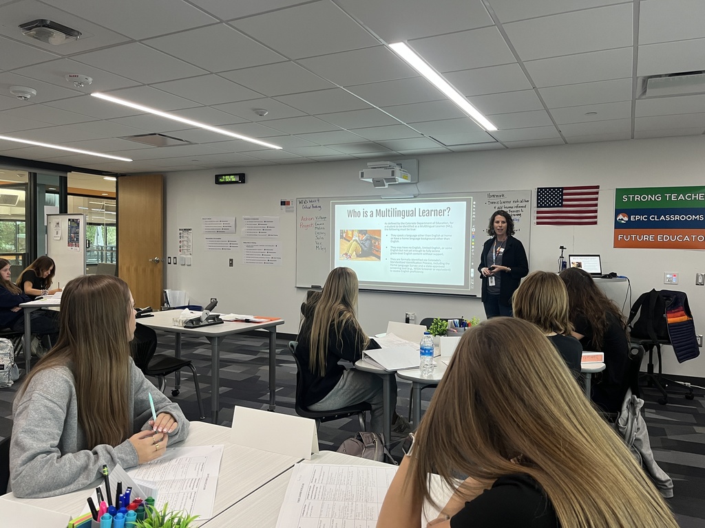 A high school classroom with students seated at white tables, actively engaging in a lesson. A female teacher stands at the front of the room, presenting a slide titled “Who is a Multilingual Learner?” A U.S. flag is mounted on the wall, along with colorful banners that read “STRONG TEACHERS,” “EPIC CLASSROOMS,” and “FUTURE EDUCATORS.” Students appear focused, with notebooks, worksheets, and writing utensils on their desks. The classroom is modern and well-lit, with a ceiling-mounted projector and digital clock above the whiteboard.