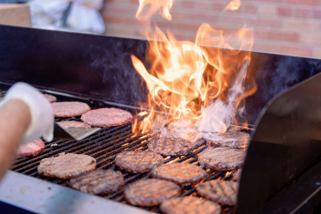 Close-up of burgers grilling on an open flame at the East Community Center Community Resource Expo, with a person using a spatula to flip them.