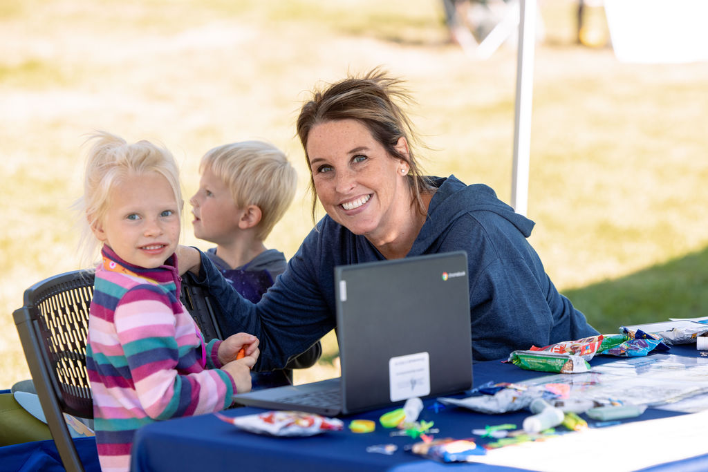 A smiling woman sits at a booth with two young children, engaging visitors with activities and information during the East Community Center Community Resource Expo.