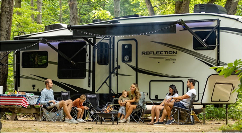 Six people of diverse ages and backgrounds sit in foldable chairs around a campfire outside a large RV labeled “Reflection” by Grand Design. The RV is parked in a wooded area with trees and green foliage. Two awnings extend from the RV, one strung with small white lights. A picnic table with a red, white, and blue American flag tablecloth is set up on the left. The group appears relaxed and cheerful, suggesting a family or friends enjoying a camping trip.