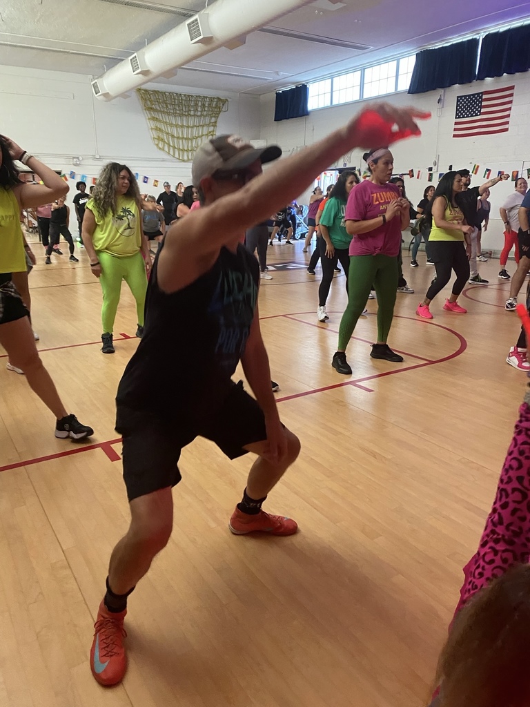A group of people in colorful workout clothes participate in a lively dance fitness class inside a gymnasium. A man in a tank top and cap is in the foreground mid-dance, surrounded by others moving and smiling as they follow along. The American flag and international flag banners hang on the wall behind them.