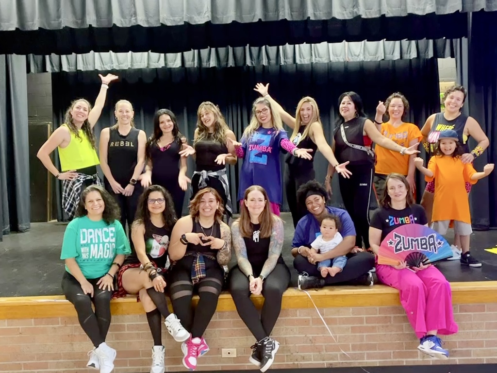 A large group of dance fitness instructors and participants pose together on and in front of a stage. Everyone is smiling and showing energetic, joyful poses. The group includes several adults and two small children. Many are wearing bright, colorful outfits with "Zumba" and dance-themed shirts.