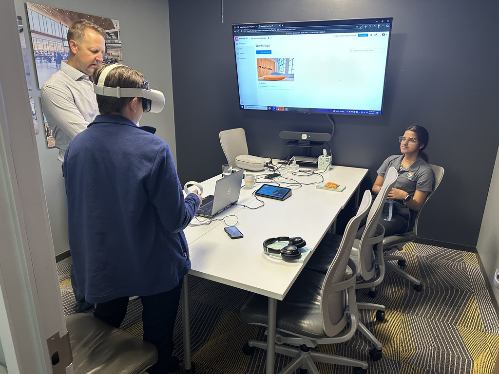 Three people in a modern conference room, with one student wearing a virtual reality headset and holding controllers. An adult male stands nearby observing, while a seated young woman watches and holds a water bottle. A laptop, tablet, phone, and headphones rest on the table, along with various cords. A large wall-mounted screen displays a webpage titled "Workshops" featuring an image of a classroom. The room has gray walls, patterned carpet, and office chairs.