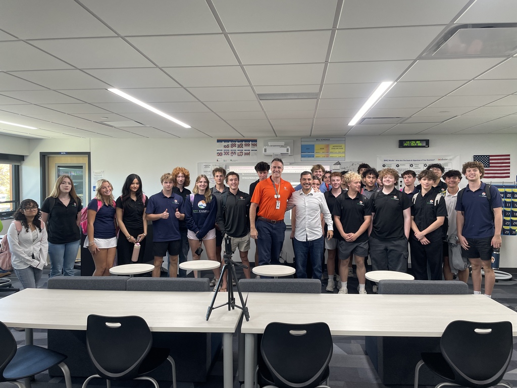 Group photo of approximately 22 high school students and two adults standing in a classroom. The group is diverse in gender and appearance, with most wearing dark-colored shirts, some featuring a mountain logo. Two adults in the center—one in an orange polo and the other in a white shirt—are smiling. Behind them are educational posters, a U.S. flag, and a digital clock showing 12:42 p.m. The room has modern furniture including tables, chairs, and a tripod set up in the foreground.