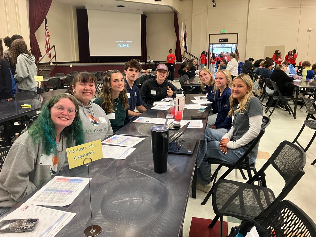 Eight high school students sit around a table in a large event space, smiling at the camera. They have name tags and worksheets in front of them, with a small sign on the table reading “Mid-Level or Experienced.” The students are diverse in appearance, with a mix of clothing including EPIC Campus apparel. In the background, many other students are seated at similar tables, and an American flag is visible on the stage. A screen at the front displays the NEC logo and the words “Empowered by Innovation.”