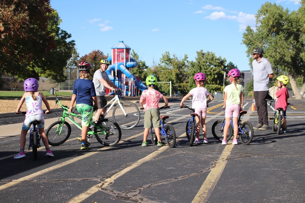 Group of children on bicycles listening to two adult instructors in a parking lot near a playground, all wearing helmets and preparing to ride.