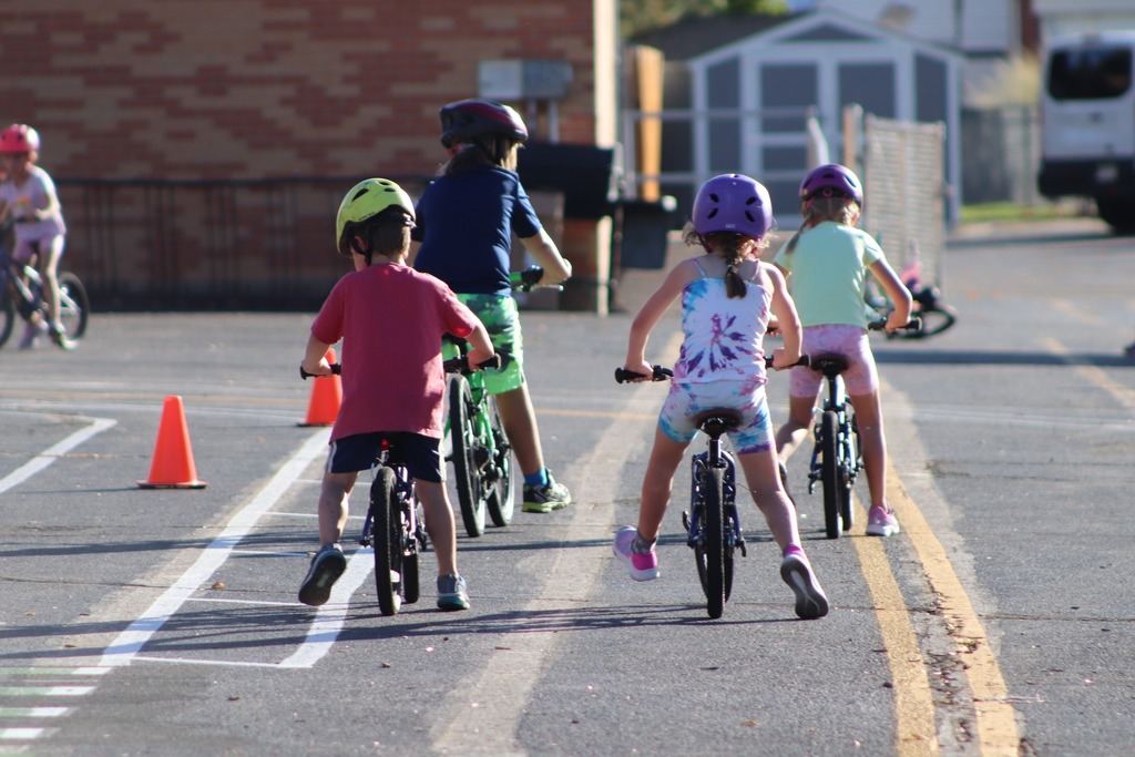 Children wearing helmets ride bicycles along a marked path with traffic lines and orange cones in a paved practice area.