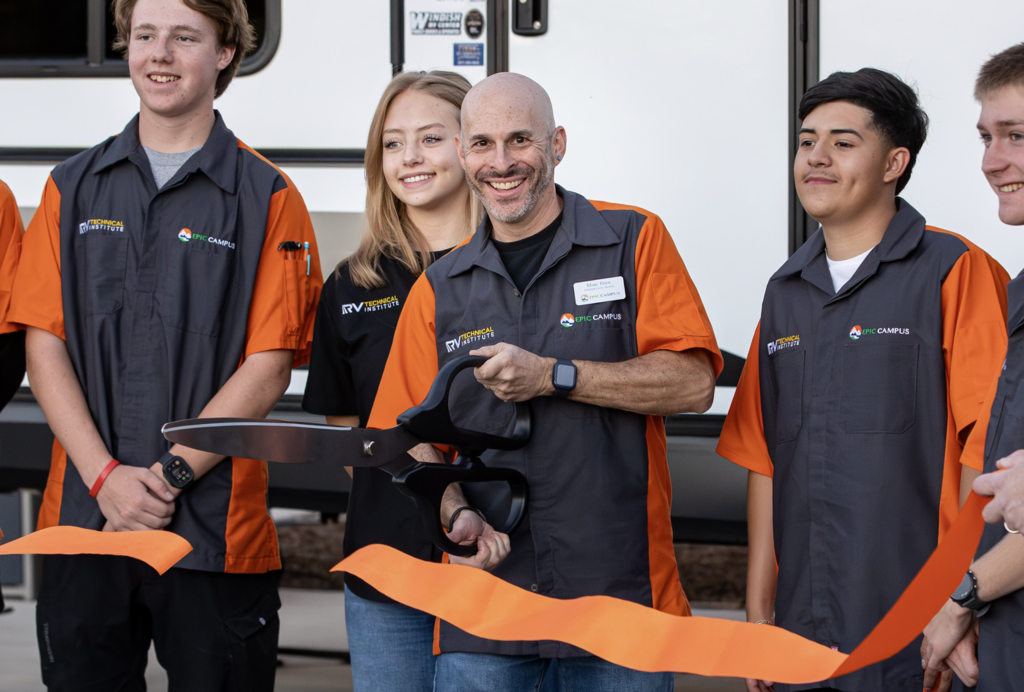 A smiling man in a grey and orange uniform cuts an orange ribbon with oversized scissors, flanked by three students in matching uniforms and a young woman.