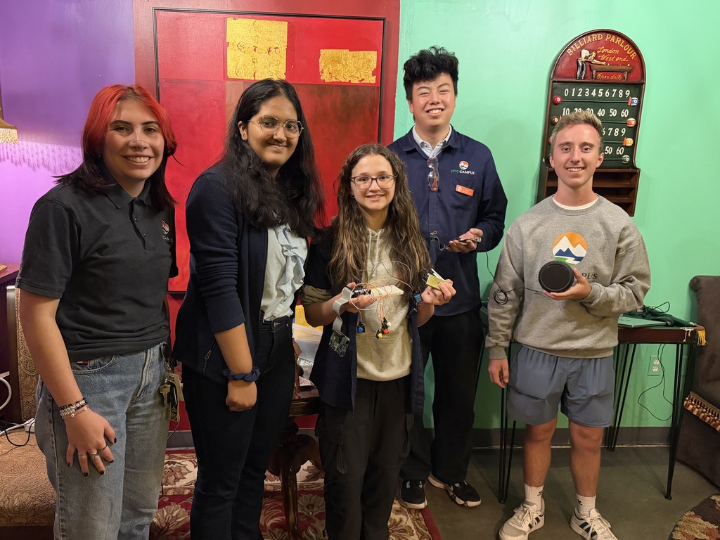 Five high school students of diverse backgrounds stand smiling indoors in a colorfully decorated room with purple, teal, and red walls. They hold small electronic devices and circuit boards, suggesting a STEM project. The student in the center wears glasses and a hoodie, holding a breadboard with wires. A “Billiard Parlour” scoreboard hangs on the wall behind them. The student on the far right holds a round black speaker. A table with a patterned rug and chairs is partially visible.
