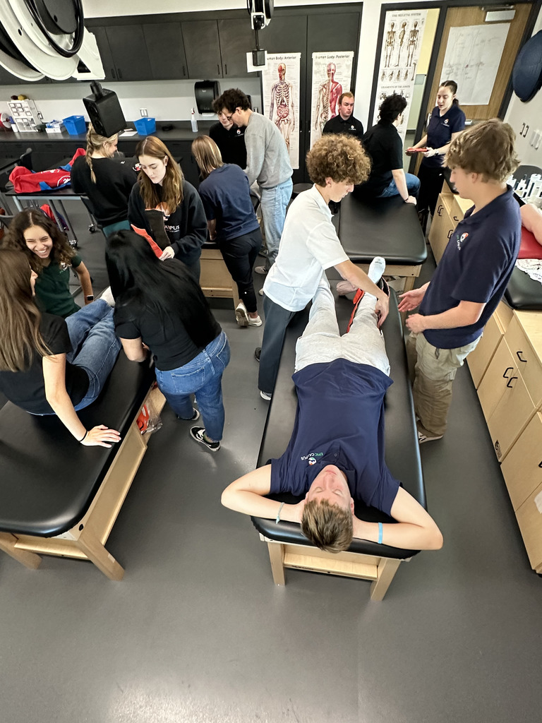 High school students practice sports medicine skills in a classroom with treatment tables. One student lies on a table while others measure and wrap his leg with athletic tape. Groups of students work in pairs or small teams, engaged in hands-on learning. Anatomical posters showing the muscular and skeletal systems are displayed on the back wall. The environment is clinical and educational, with medical supplies, cabinets, and charts visible throughout the room.