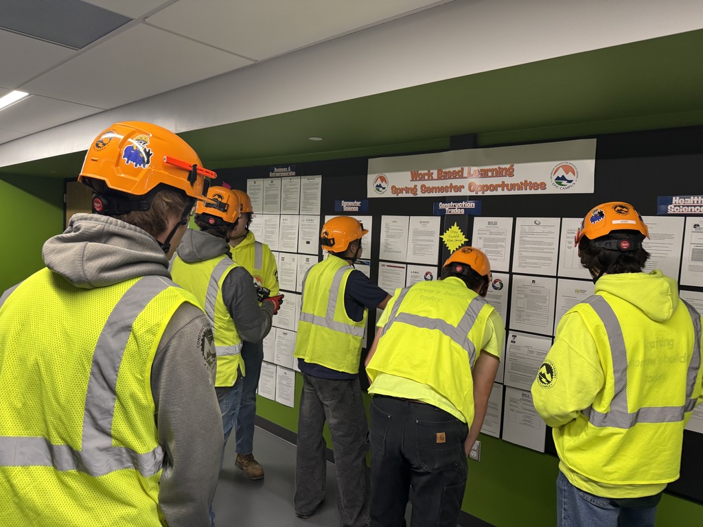 A group of high school students wearing neon yellow safety vests and bright orange construction helmets gather in front of a bulletin board titled “Work Based Learning Spring Semester Opportunities.” The students, seen mostly from behind, appear to be reading posted internship and job listings under categories like Business, Construction, and Health Sciences. The setting is an indoor hallway with green and gray walls, suggesting a career and technical education environment. Some hard hats are decorated with stickers and logos.