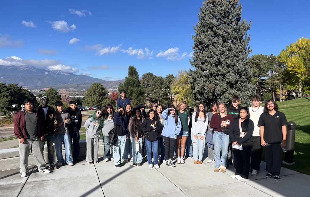 A diverse group of high school students stands together outdoors on a sunny day, posing for a group photo on a paved path. They are surrounded by green grass, autumn-colored trees, and a large evergreen tree. In the background, mountains rise under a bright blue sky with scattered clouds. Most students wear casual clothing and a few are in school-branded sweatshirts. Some are smiling while others squint or shade their eyes from the sun.