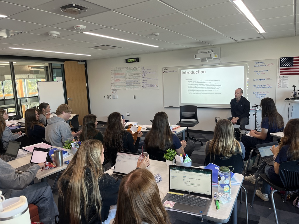 A classroom full of high school students listens attentively to a male guest speaker seated at the front. The speaker presents a slide titled “Introduction” projected onto a whiteboard, while notes and posters are visible on nearby walls, including an American flag. Students are seated at tables with laptops, notebooks, and drinks. The room has modern furniture and large windows letting in natural light. A camera tripod and mic setup suggests the session may be recorded or livestreamed.