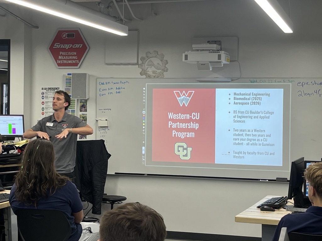 A guest speaker from Western Colorado University stands in front of a high school classroom, presenting a slide titled “Western-CU Partnership Program.” The slide lists degree options including Mechanical Engineering, Biomedical (2025), and Aerospace (2026), with details about a dual enrollment program with CU Boulder. Students sit at desks with computers, listening attentively. A “Snap-on Precision Measuring Instruments” sign and various STEM-related posters are visible on the walls.