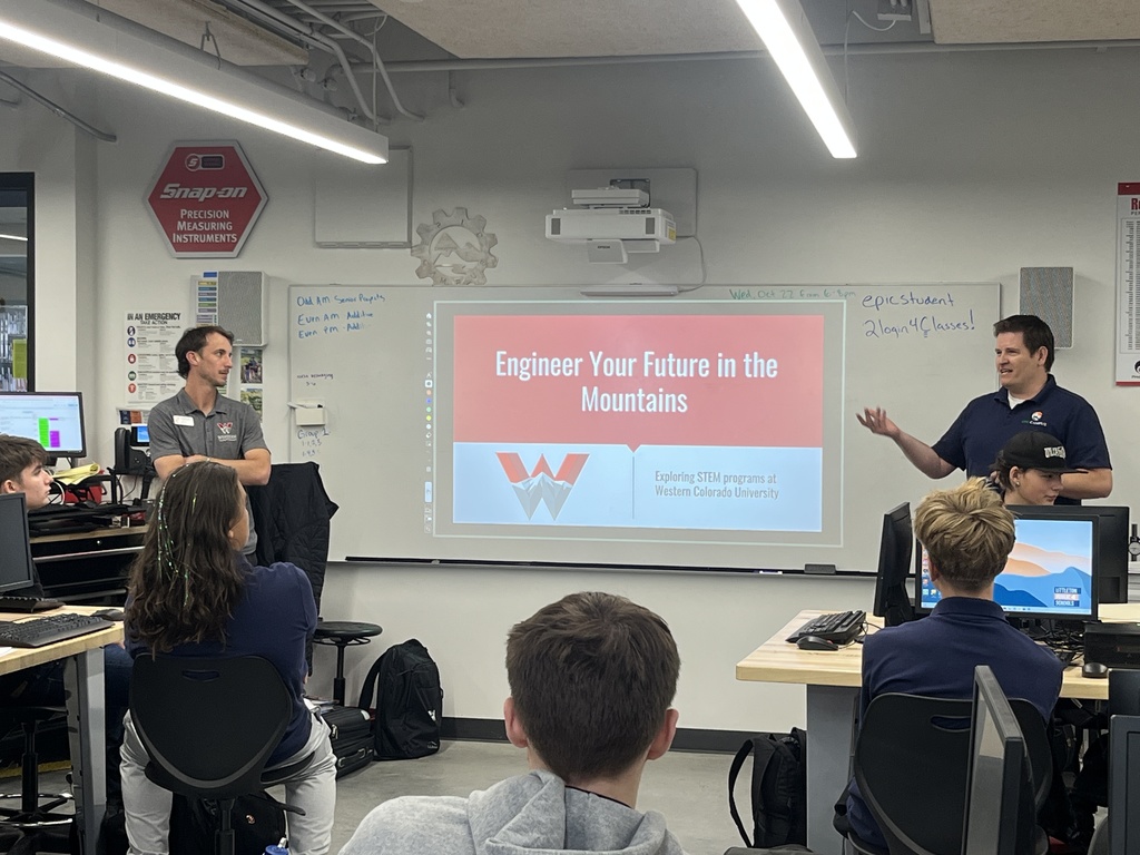 Two presenters from Western Colorado University speak to high school students in a computer lab. A projected slide reads “Engineer Your Future in the Mountains – Exploring STEM programs at Western Colorado University.” One speaker gestures as he talks, while the other listens. Students face the presenters with computers in front of them. The classroom is equipped with technical lab furniture and STEM posters on the walls.
