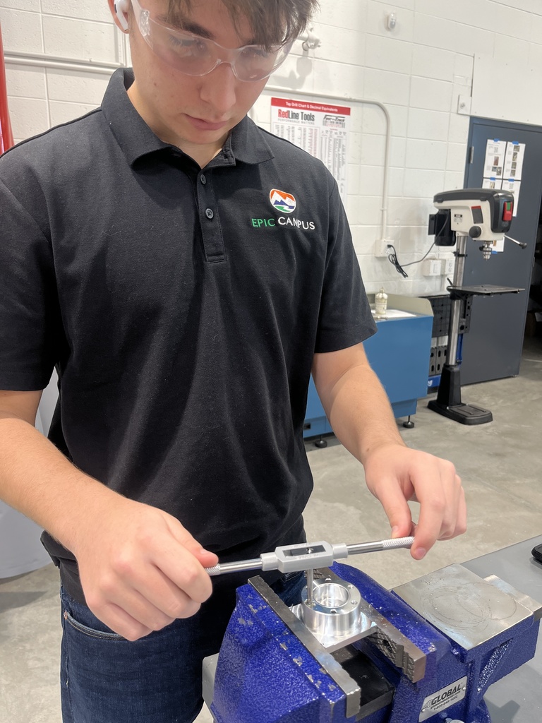 A high school student wearing safety goggles and an EPIC Campus polo shirt uses a tap wrench to thread a metal part held in a blue vise. The student is working in a well-lit workshop with white walls, industrial equipment, and a drill press visible in the background. A safety chart titled “RedLine Tools” hangs on the wall.