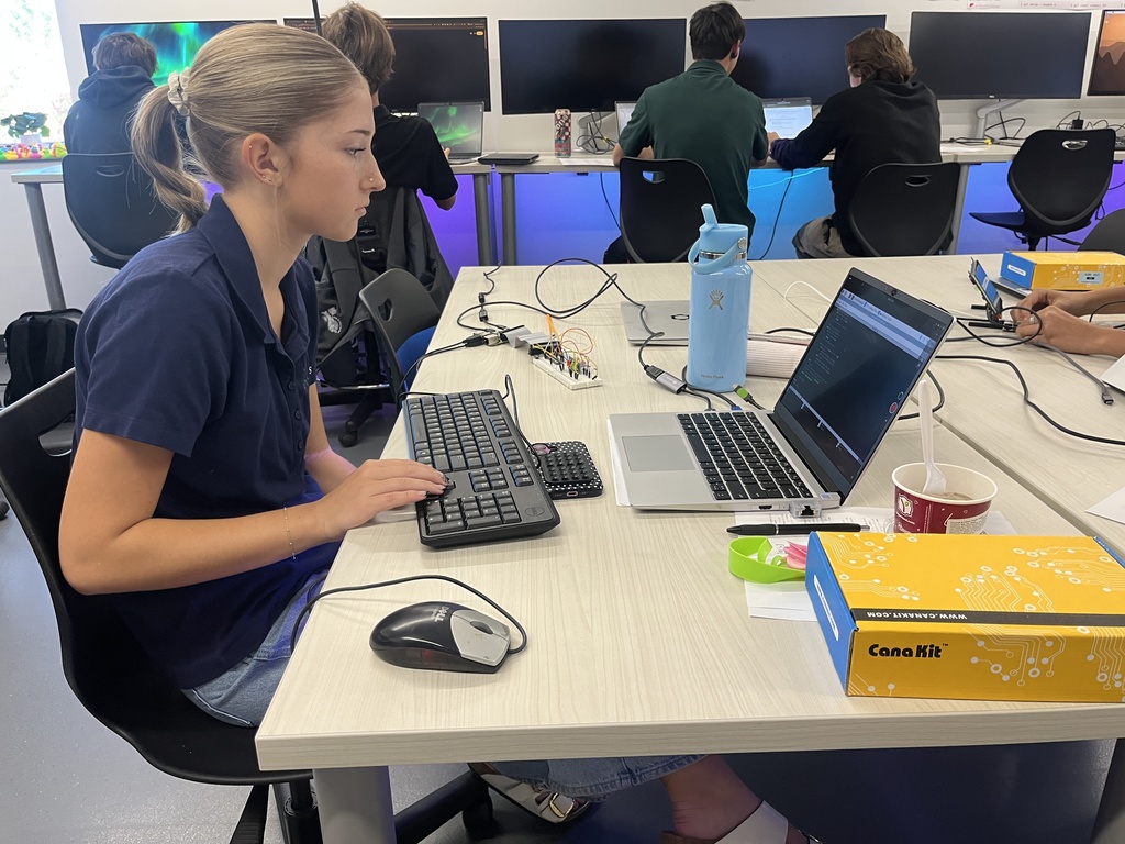 A high school student works at a desk with a laptop and external keyboard, writing code on the screen. A small breadboard with wires and electronic components is connected nearby. A yellow CanaKit box and a blue water bottle sit on the table. In the background, other students work at computers with colorful LED lighting under the desk.
