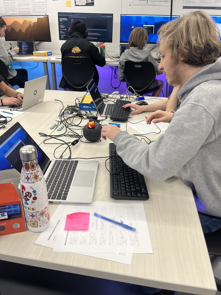 A student in a gray hoodie sits at a table with a laptop, keyboard, and a connected electronic project. A small rubber duck sits on a round speaker device. The table has scattered materials, including notes, a colorful water bottle, and a red electronics box. Several students in the background are coding on desktop monitors in a tech lab setting with ambient purple lighting.