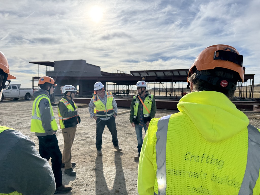 A group of construction workers and supervisors wearing safety vests and hard hats stand in a circle at a construction site under bright daylight. In the background, steel beams and framing of a building are partially erected. The person in the foreground wears a neon yellow vest with partially visible text reading "Crafting Tomorrow’s Builders Today." The group includes individuals of diverse backgrounds, and the setting appears collaborative and instructional.