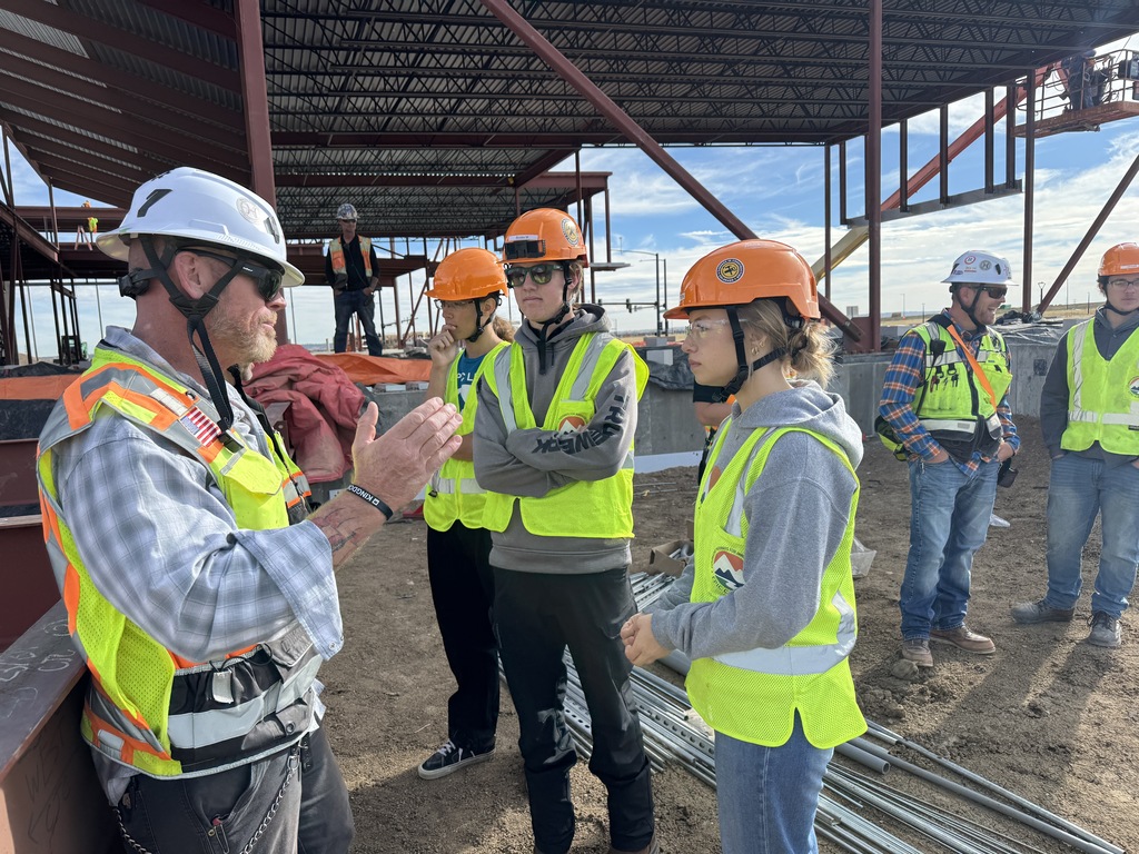 A construction supervisor wearing a white hard hat and safety vest gestures while speaking to a group of students or young trainees at a building site. The group, dressed in bright safety vests and hard hats, listens attentively near steel framing and building materials. A large structure with exposed beams and girders is visible in the background. The group represents a mix of genders and backgrounds, highlighting a hands-on learning environment.