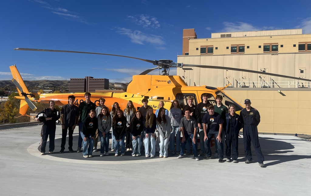 A group of high school students stands in a semi-circle around an orange AirLife medical helicopter on a rooftop helipad. Two flight crew members in navy uniforms are speaking to the students, one holding medical equipment near the open side door of the helicopter. The students, dressed casually in jeans and sweatshirts, listen attentively under a partly cloudy sky.