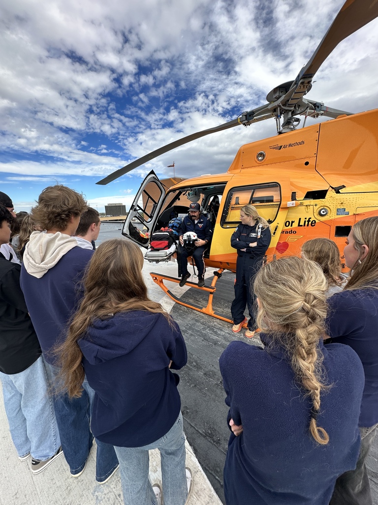 A large group of high school students poses in front of a bright orange AirLife medical helicopter on a rooftop helipad. They are joined by two flight crew members in navy uniforms. Most students wear matching EC@CAMPUS or LPS-branded shirts, and the city skyline and distant hills are visible in the background under a clear blue sky.