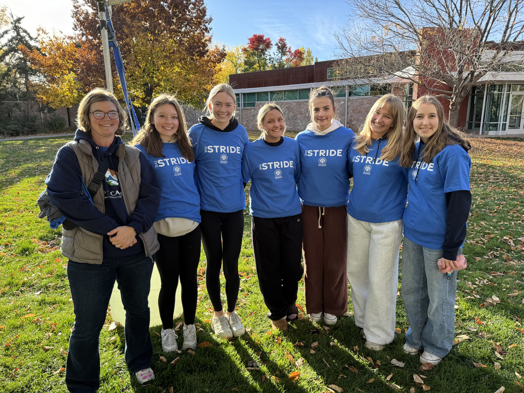 Seven women stand in a row on a grassy area with autumn leaves and trees in the background. Six of them wear matching blue "STRIDE" event T-shirts, while one adult on the far left wears an EC@CAMPUS hoodie and vest. They are smiling and appear to be participating in or volunteering at a community or school event. A building with large windows and red-brown brick is visible behind them.