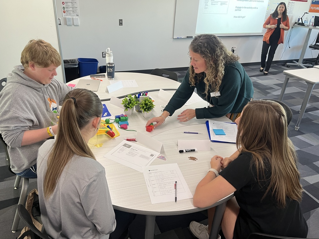 A group of three students sits at a round table working with colorful geometric blocks and worksheets, while a woman in a green sweater leans over to assist. Another adult stands near a whiteboard in the background, where a presentation is projected. The classroom has modern furniture and a gray patterned floor. The students appear engaged in a hands-on learning activity.