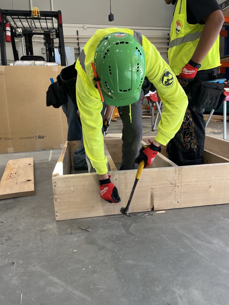 A student in a bright green hard hat and neon safety hoodie uses a hammer to remove nails from a wooden frame on the floor of a workshop. Another student in a yellow safety vest and gloves stands nearby. Both are wearing tool belts, and construction tools and materials are visible around the workspace.