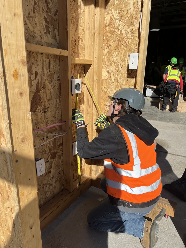 A student in a gray hard hat and orange reflective vest kneels while measuring and working on electrical wiring within a framed wooden wall structure. The student uses a measuring tape to mark a spot near an installed outlet. Other individuals in safety gear are visible in the background, suggesting a construction training environment.