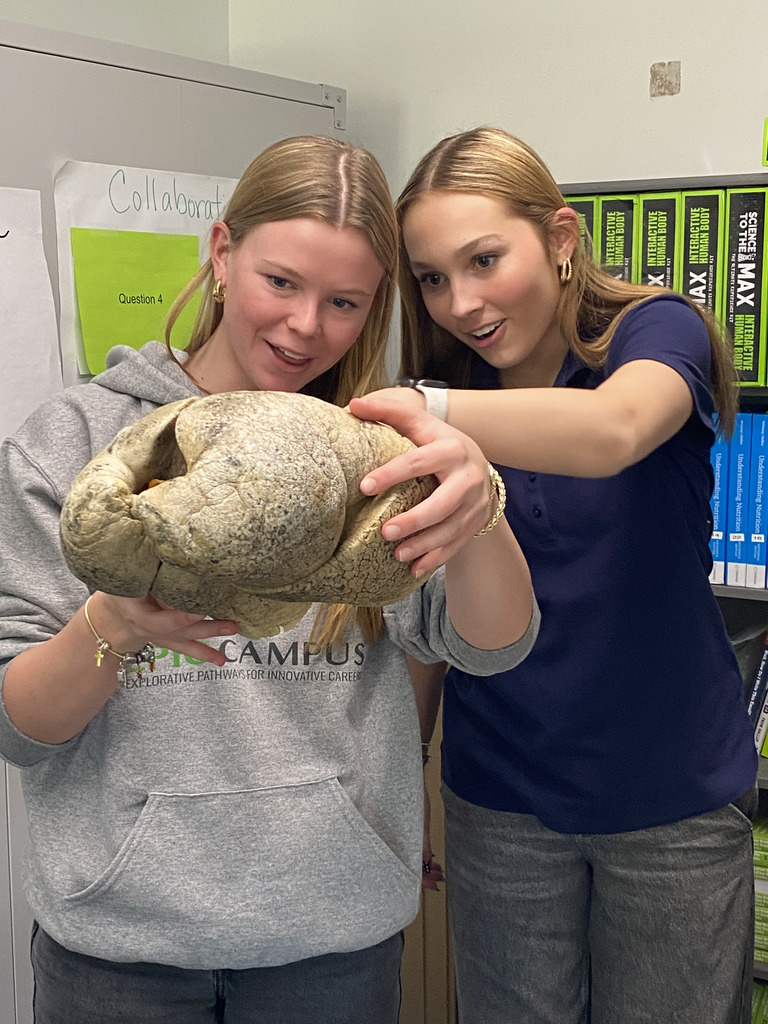 Two high school students excitedly examine and hold a large, textured object that appears to be a fossil or replica of a skull. One student wears an EC@CAMPUS hoodie, and the other is dressed in a navy polo. Behind them are classroom shelves with science books and posters, suggesting a hands-on learning activity in a science or career-exploration setting.