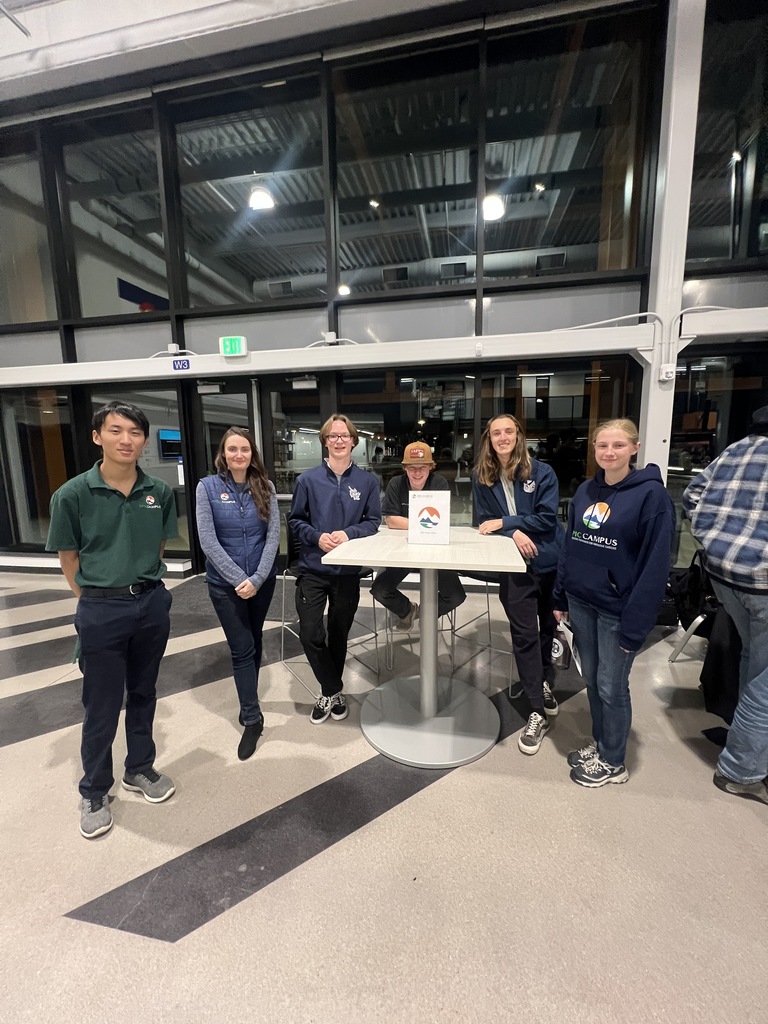 Group of six young people, a mix of students and staff, smiling and posing in front of a round table with a printed sign. They are standing in a well-lit, glass-walled area with high ceilings. Most are wearing sweatshirts with school or program logos.