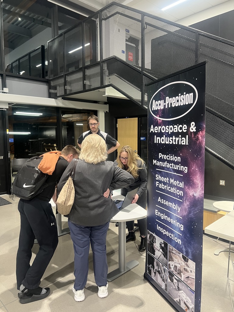 Close-up of a booth for “Accu-Precision Aerospace & Industrial,” featuring a large vertical banner listing services such as precision manufacturing and sheet metal fabrication. Two women behind the table assist attendees. A staircase and modern interior architecture are visible in the background.