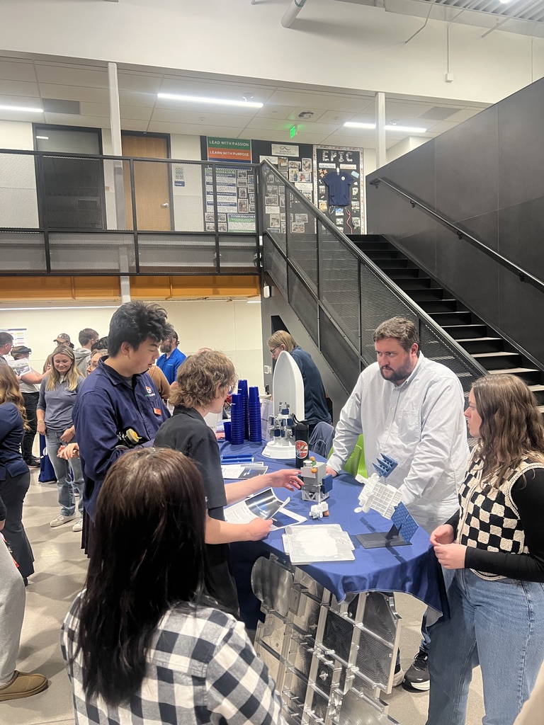 Students and adults gather around a booth displaying small models of spacecraft and satellites on a blue tablecloth. A man and woman explain items on the table while students hold informational materials. A staircase and bulletin board labeled “EPIC” are visible in the background.