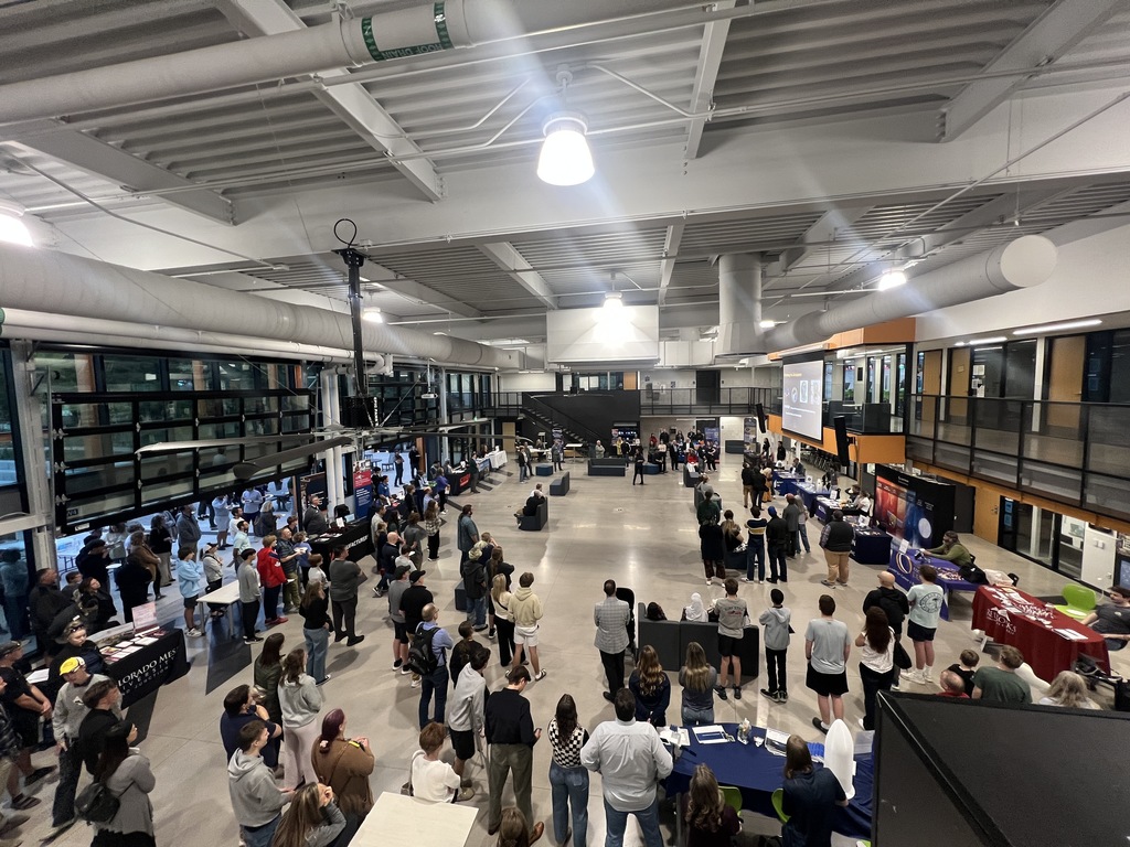 Wide-angle view of a bustling indoor event in a modern, industrial-style space with high ceilings and exposed ducts. Multiple vendor tables line the perimeter, while attendees—students, families, and professionals—interact and explore. A large screen displays visuals on one wall, and branding materials are visible at several tables.