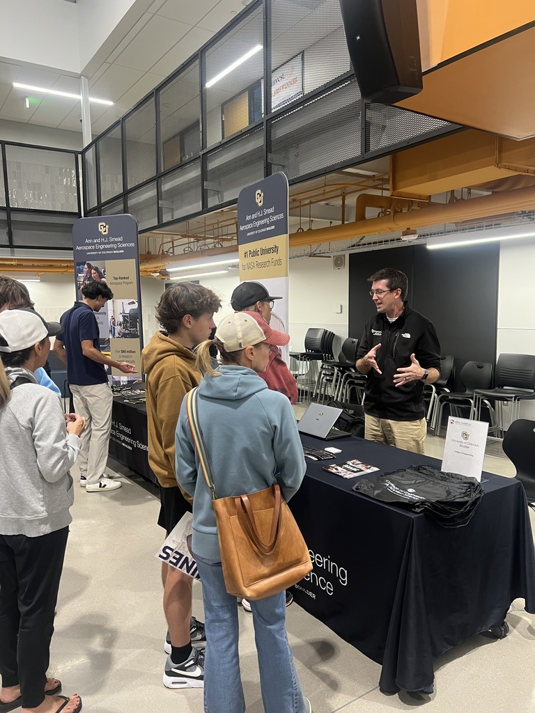 University of Colorado Aerospace Engineering booth with a representative engaging a group of students and parents. Two vertical banners advertise CU’s national ranking and NASA research funding. The booth has brochures, swag, and a laptop on a black tablecloth.
