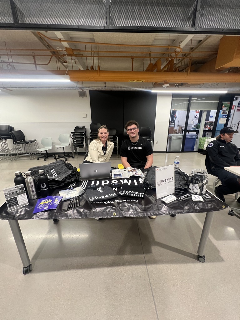 Two Upswing Foundation representatives smile from behind a table featuring merchandise including water bottles, brochures, and drawstring bags. The black tablecloth is branded with the Upswing Foundation logo. Empty chairs and classroom spaces are visible in the background.
