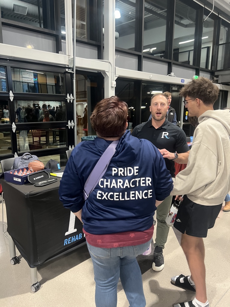 A man from Rehab United Physical Therapy & Sports Performance talks with two attendees at a booth. One attendee wears a navy hoodie with the words "PRIDE CHARACTER EXCELLENCE" printed in white on the back. Branded items and a football are displayed on the black table with a Rehab United logo.
