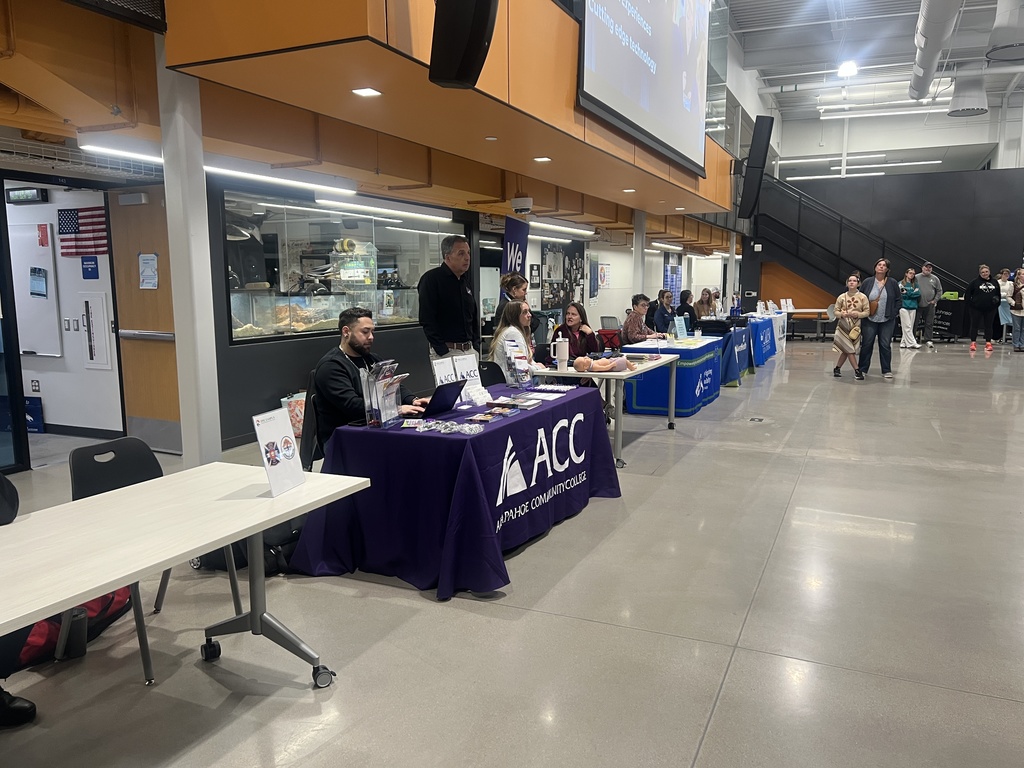 A row of booths with representatives seated at tables from various institutions, including Arapahoe Community College (ACC), line a hallway. ACC’s purple tablecloth is prominently visible. Attendees interact with representatives, while others observe nearby.