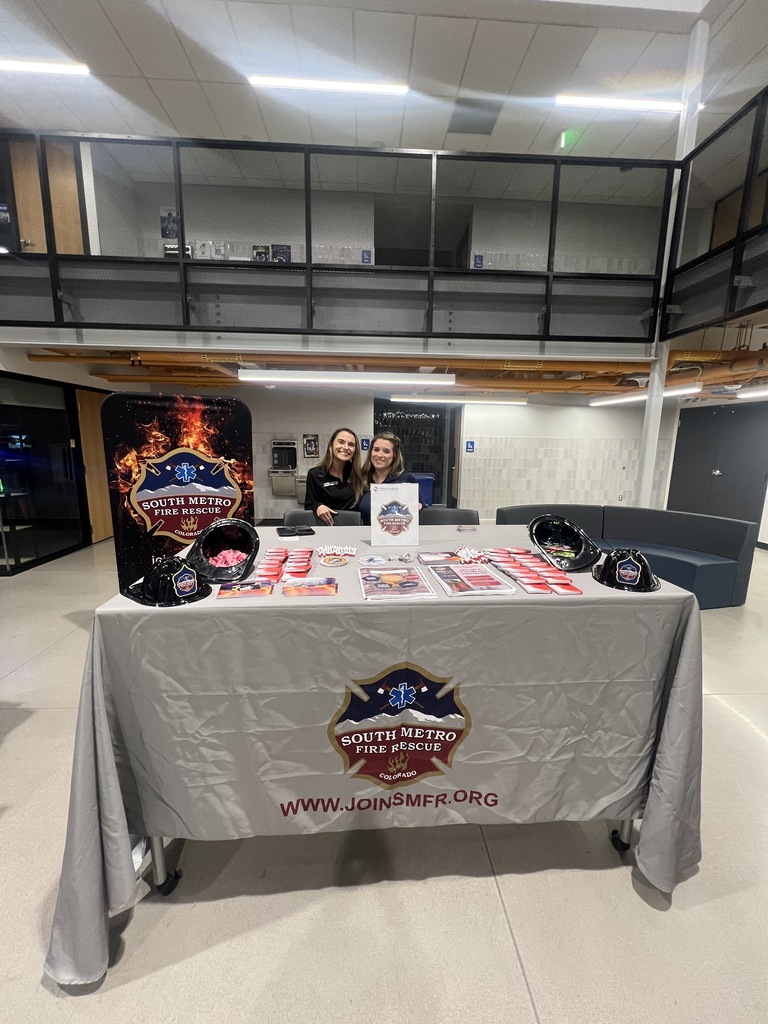 Two smiling representatives from South Metro Fire Rescue stand behind a booth with a gray tablecloth displaying the department’s logo. Promotional items such as red buttons and toy helmets are neatly arranged. A vertical banner and a fire department flyer holder stand beside the table.