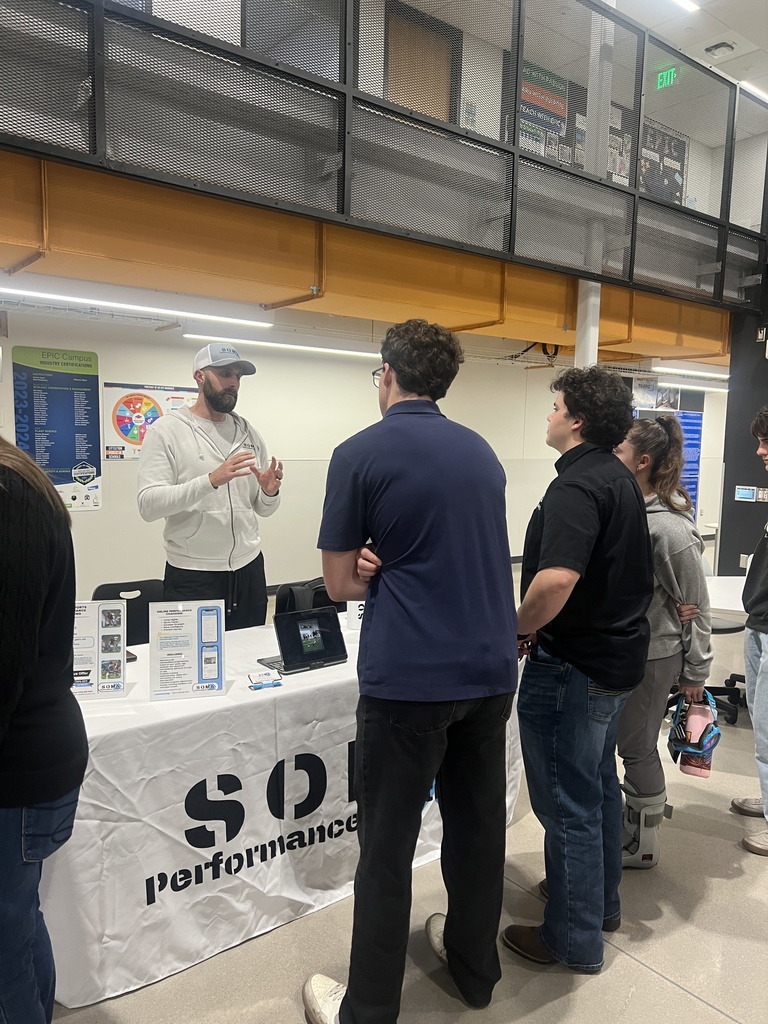 A man in a white hoodie and baseball cap speaks to a small group of students at a booth labeled “SOM Performance.” The table has information displays and a tablet showing a performance video. The background includes posters and classroom doors.