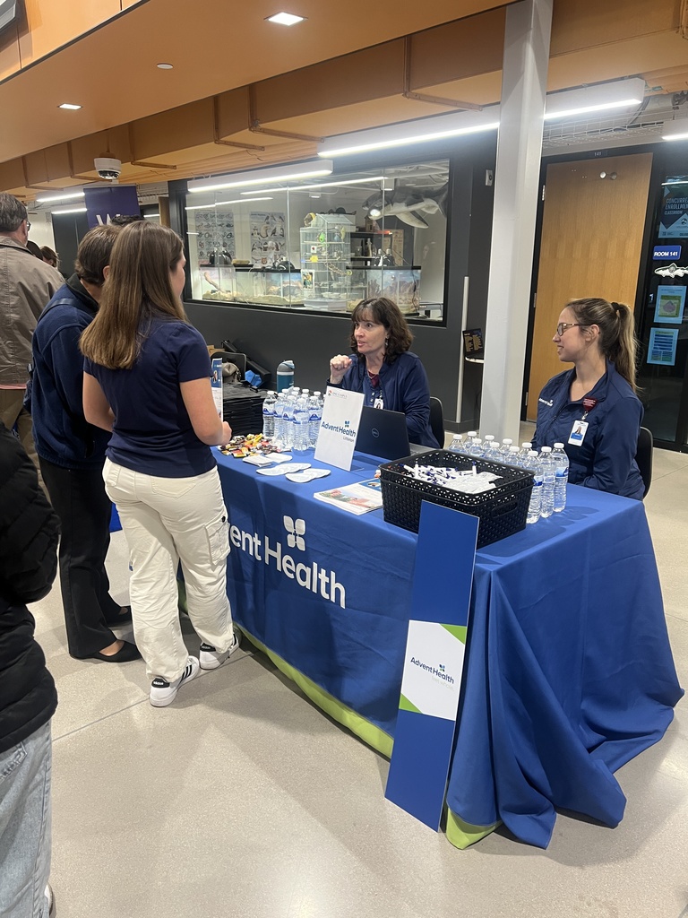 Two women from AdventHealth engage with students at a table covered in a blue branded tablecloth. The table has a laptop, brochures, snacks, and water bottles. Behind them is a glass display case featuring anatomical and biological models.