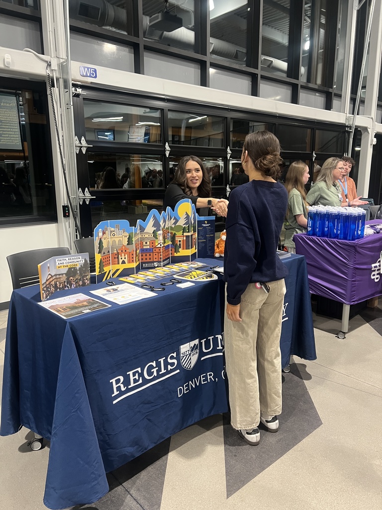 A Regis University representative smiles and shakes hands with a student at a table covered in a navy blue tablecloth with the university’s logo. The table features colorful paper cutouts of campus scenes, brochures, and orange giveaways. Two more women are seated at the adjacent booth to the right.