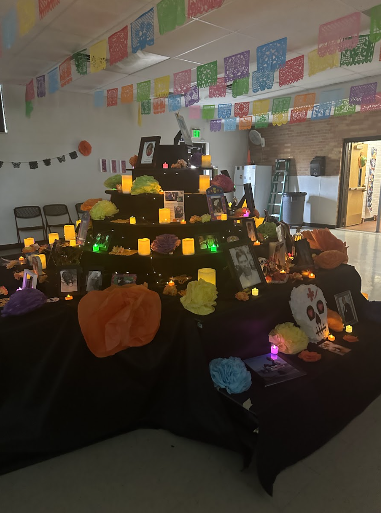 A large multi-tiered Día de los Muertos altar covered in black fabric, decorated with bright paper flowers, candles, and framed photos of loved ones. Colorful papel picado banners hang from the ceiling, and the tiered display glows with warm lights and offerings.